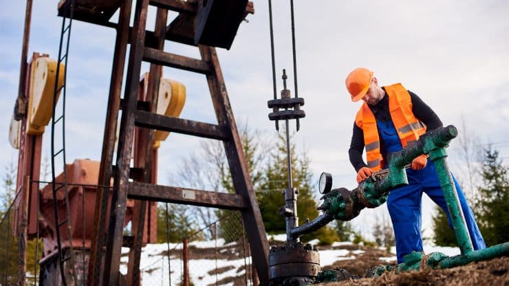 Oil worker in orange uniform and helmet working with a pipe wrench near an oil pump jack